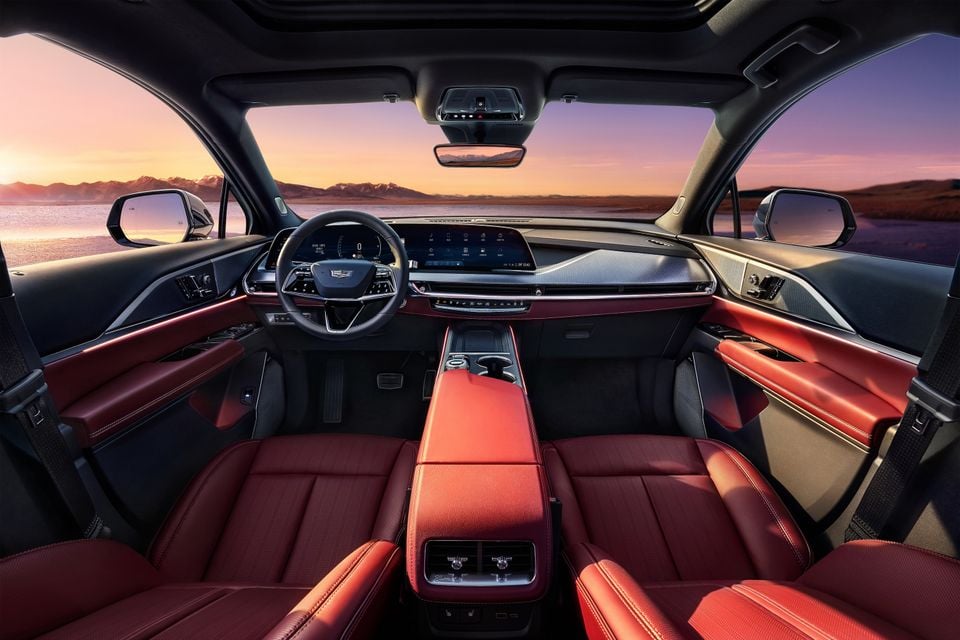 Interior of a 2024 Cadillac XT5 showing red leather seats, large dual infotainment screens, and a centre console with a panoramic sunset view through the windscreen