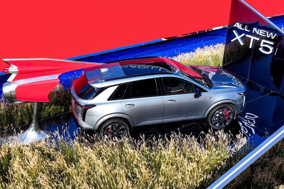 A silver Cadillac XT5 SUV seen from the side at a promotional display, surrounded by grass and bold red and blue mirrored backdrops with 'All New XT5' branding.