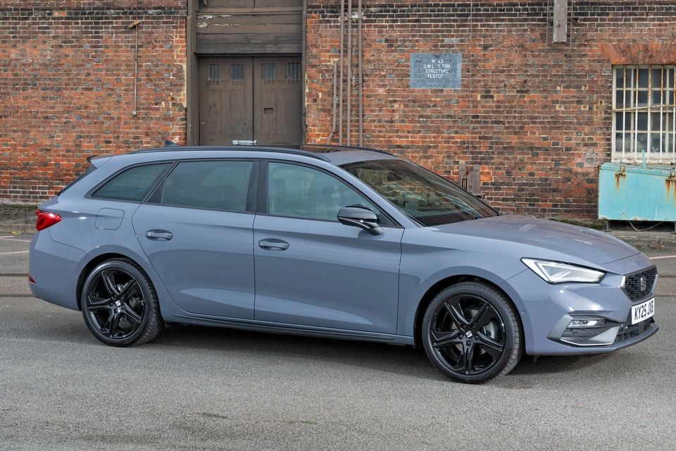 A grey 2025 SEAT Leon wagon seen from the side, parked in front of a brick industrial building