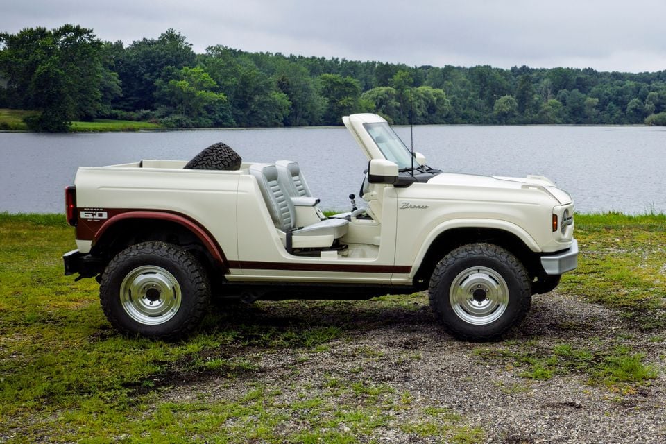 A cream Ford Bronco Roadster concept with no roof or doors, seen from the side, parked near a lake with trees in the background