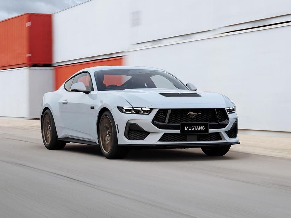 A white Ford Mustang coupe seen from the front-side angle, driving along a road with a blurred background.
