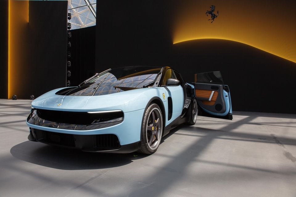 A light blue Ferrari Luce supercar with its door open, seen from the front three-quarter angle at an indoor reveal event, with the Ferrari prancing horse logo visible in the background.