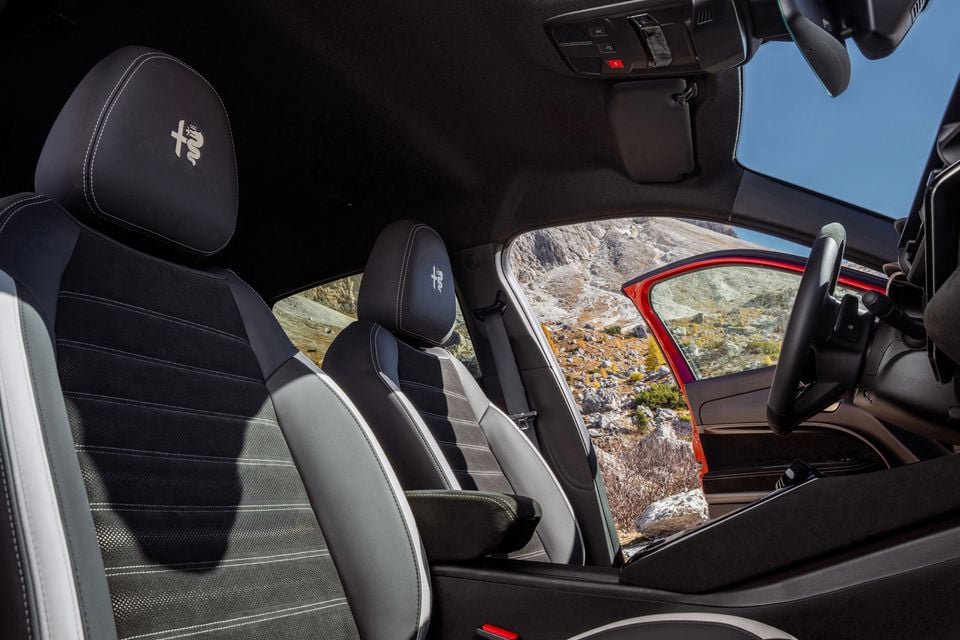 Interior of a 2026 Alfa Romeo Junior Sport Speciale showing front seats with Alfa Romeo logo embroidered headrests, contrasting grey and black upholstery with white stitching, and a panoramic sunroof overlooking a rocky mountain landscape