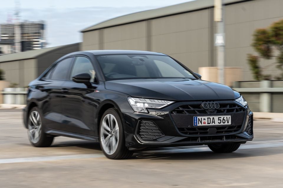 A black 2026 Audi A3 sedan S-line driving through an urban area, seen from the front-left side