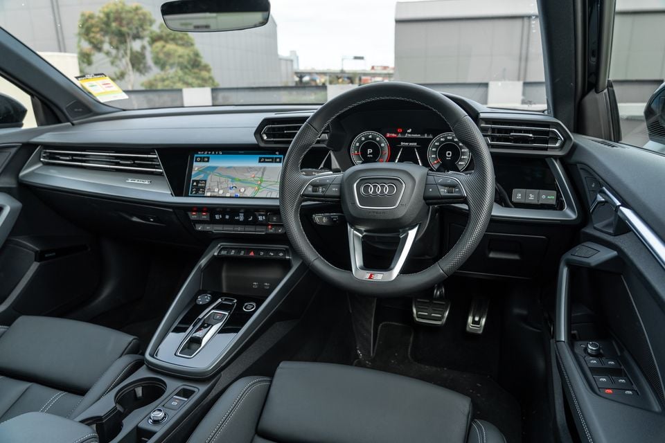 Interior of a 2026 Audi A3 S line sedan showing the leather-trimmed steering wheel with S badge, digital instrument cluster, touchscreen infotainment with navigation, and centre console with automatic gear selector