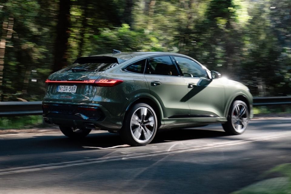 A green 2026 Audi Q5 e hybrid Sportback seen from the rear three-quarter angle while driving through a tree-lined road