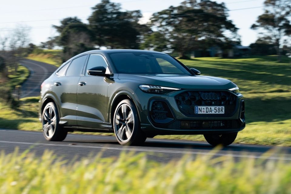 A dark green 2026 Audi Q5 Sportback SUV-coupe driving on a country road, seen from the front-side