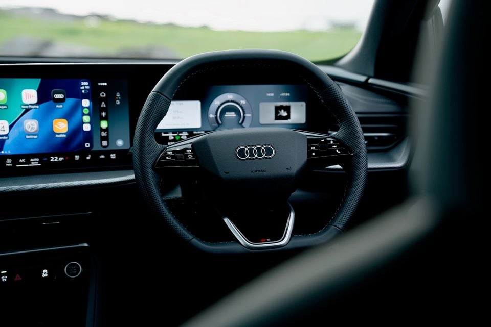 Interior of a 2026 Audi Q5 e-hybrid showing the leather-trimmed steering wheel with Audi four-rings logo, digital instrument cluster, and infotainment touchscreen displaying Apple CarPlay.