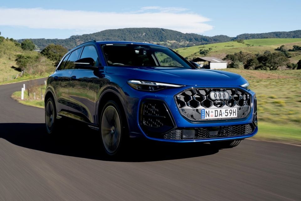 A blue 2026 Audi Q5 SUV driving on a rural road, seen from the front-side angle