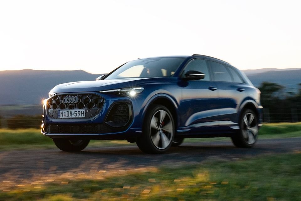 A blue 2026 Audi Q5 e hybrid SUV seen from the front-side while driving on a rural road at dusk