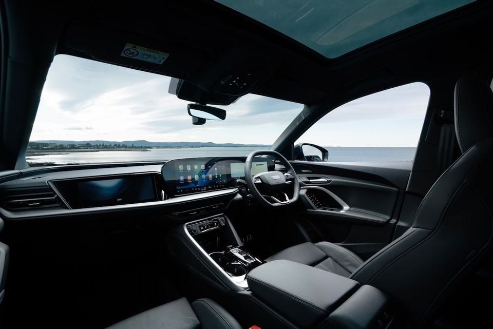Interior of a 2026 Audi Q5 e hybrid SUV showing the dashboard, dual touchscreens, steering wheel, and dark leather seats, with a coastal water view through the windscreen