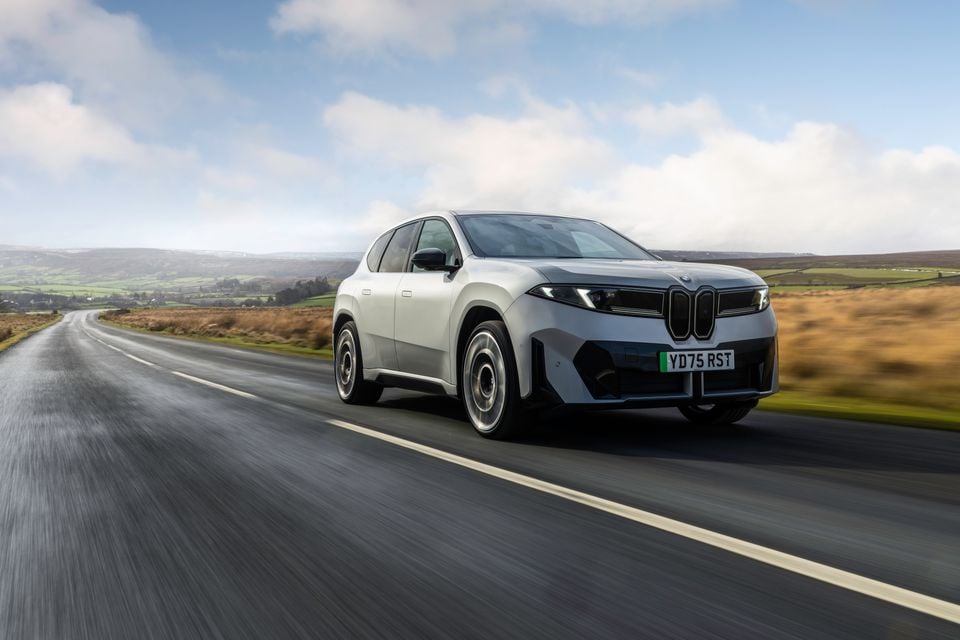 A white 2026 BMW iX3 SUV driving along a rural road, seen from the front-side angle