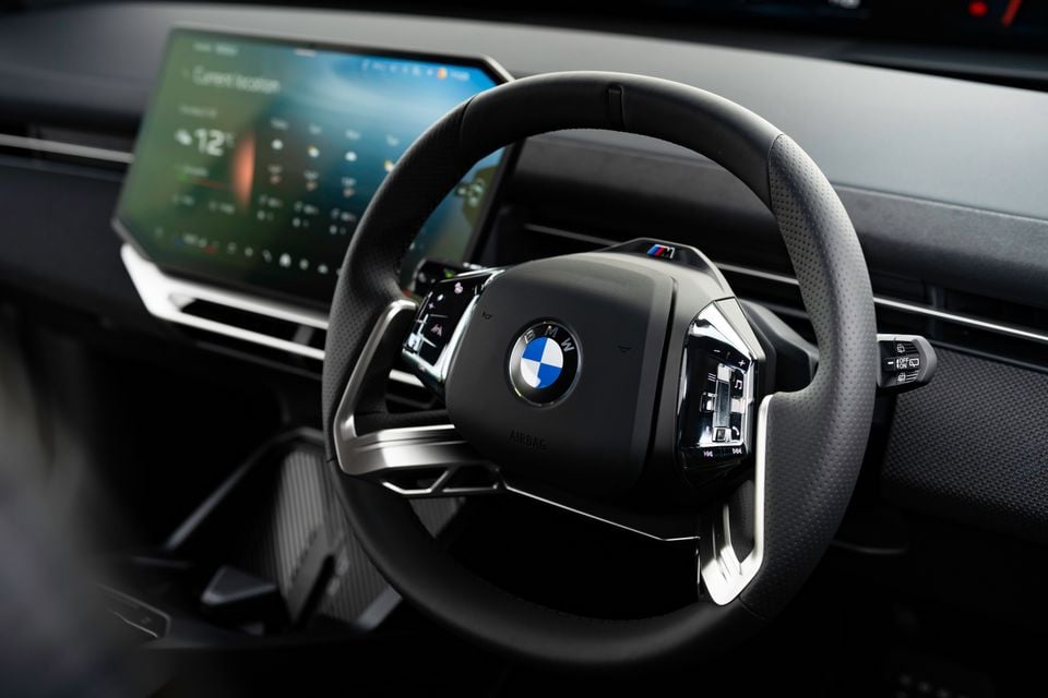 BMW M Sport steering wheel with iDrive controls and a large curved infotainment touchscreen visible in the background, inside a 2026 BMW iX3.