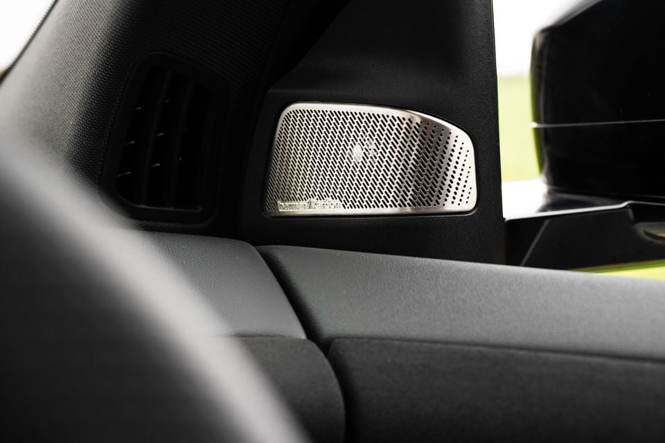 A Harman Kardon speaker mounted on the dashboard of a 2026 BMW iX3, with green exterior visible through the window
