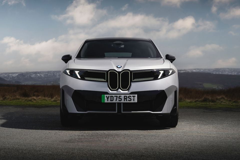 A silver BMW iX3 SUV seen from the front, parked on a rural road with a cloudy sky and hills in the background.