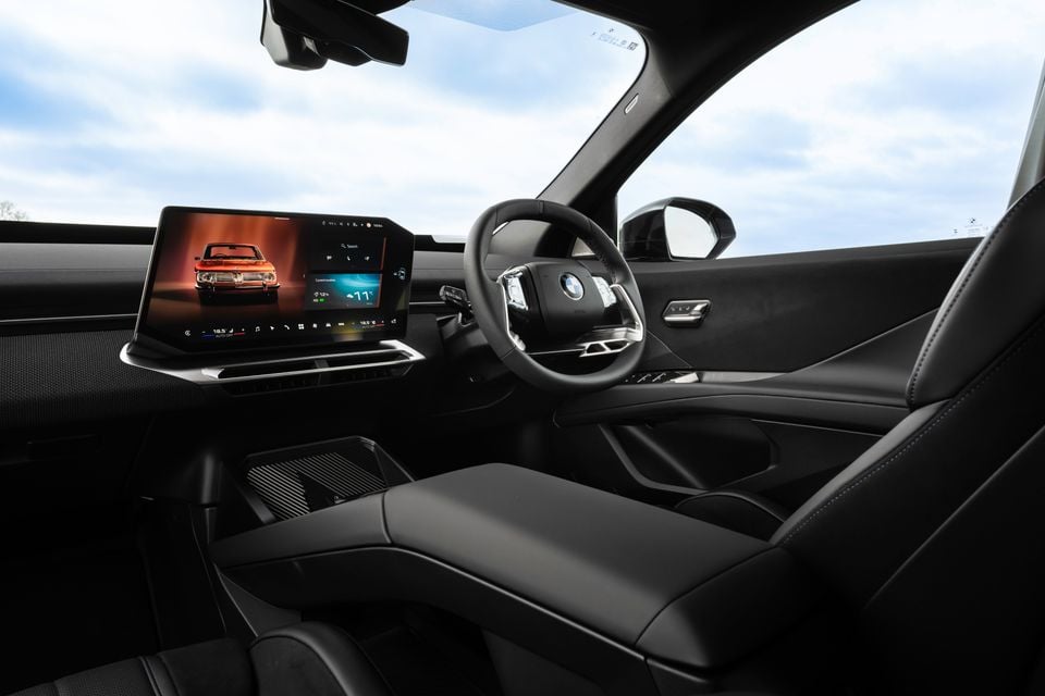 Interior of a BMW iX3 showing the dashboard, curved infotainment touchscreen, steering wheel, and black leather seats from the passenger side
