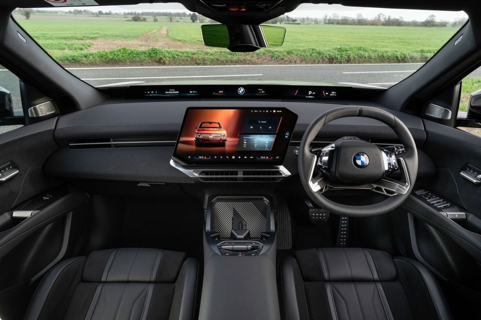 Interior of a 2026 BMW iX3 M Sport Pro showing the dashboard, large central touchscreen infotainment display, M steering wheel, and front seats, viewed from the rear
