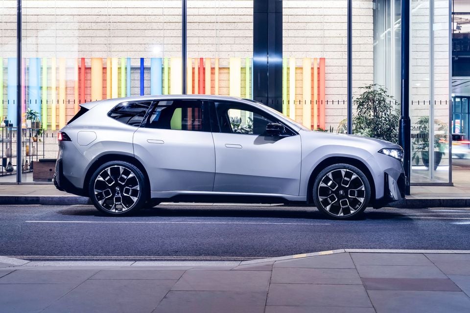A silver 2026 BMW iX3 SUV seen from the side, parked on a city street in front of a building with colourful window displays.