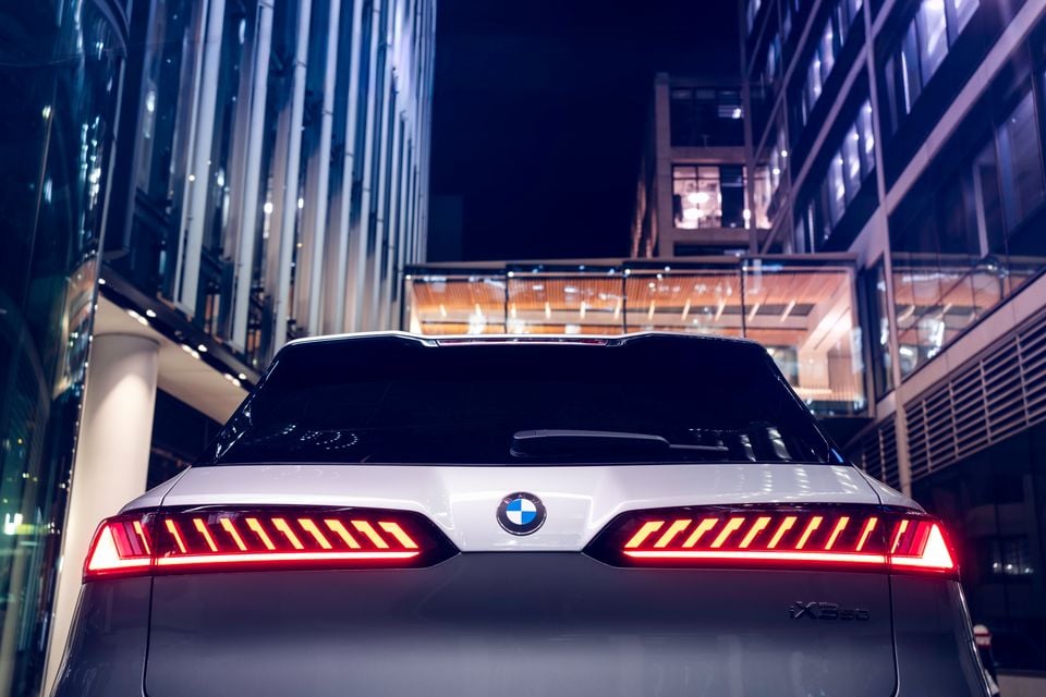 Rear of a silver BMW iX3 SUV with illuminated tail-lights, photographed at night in an urban setting with glass buildings in the background.