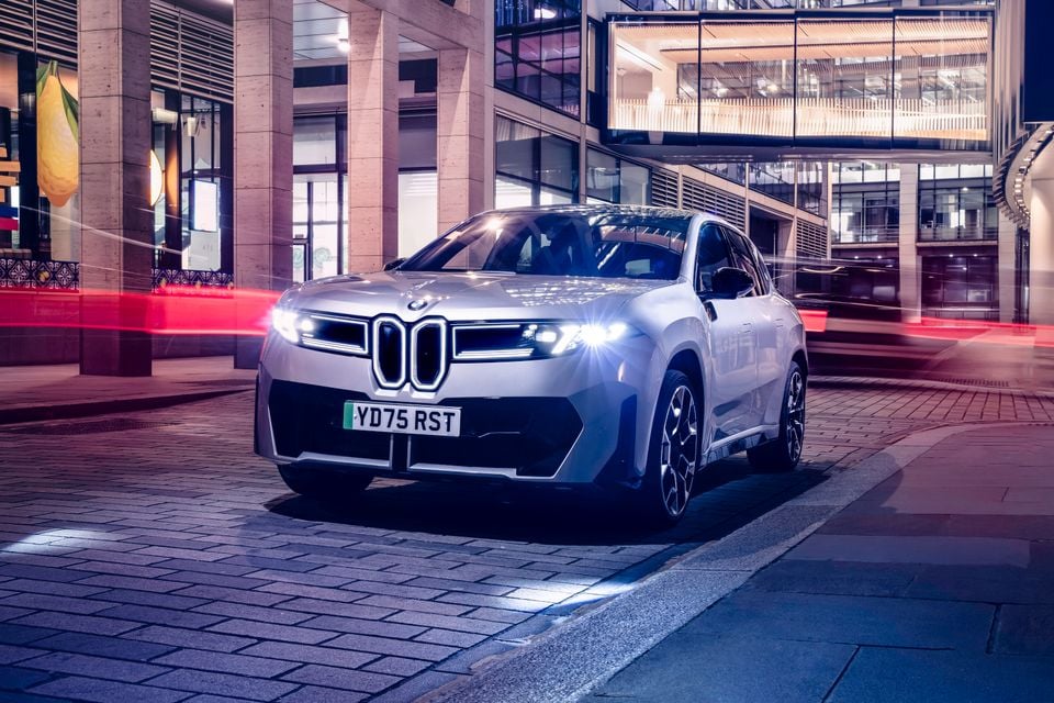 A silver 2026 BMW iX3 SUV seen from the front at night in an urban setting with its headlights on