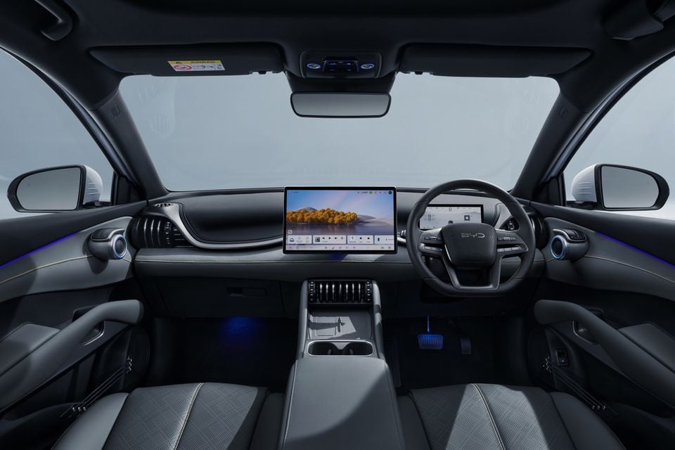 Interior of a BYD Atto 3 Evo showing the dashboard with a large central touchscreen, BYD steering wheel, ambient lighting, and grey fabric seats, seen from the rear