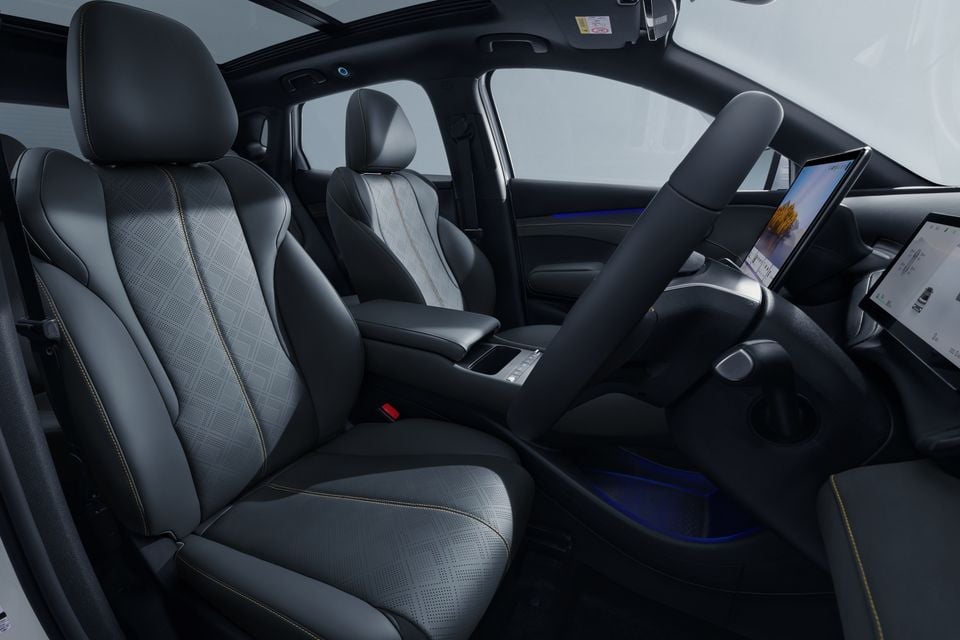 Interior of the 2026 BYD Atto 3 Evo showing the front seats with grey and black patterned upholstery, gold contrast stitching, a large central touchscreen, and ambient blue lighting