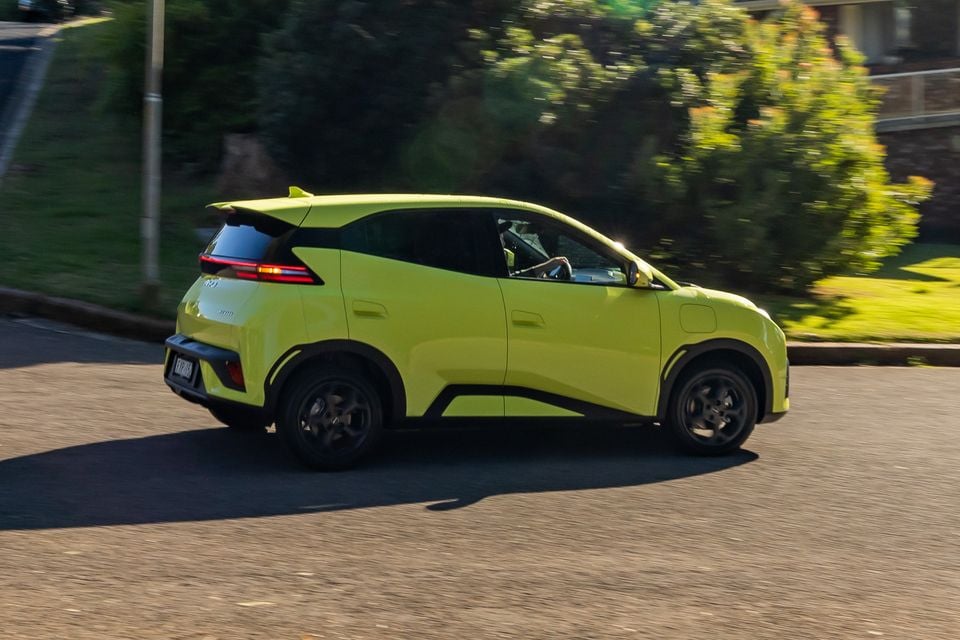A lime green 2026 BYD Atto 1 small SUV driving along a suburban street, seen from the rear side