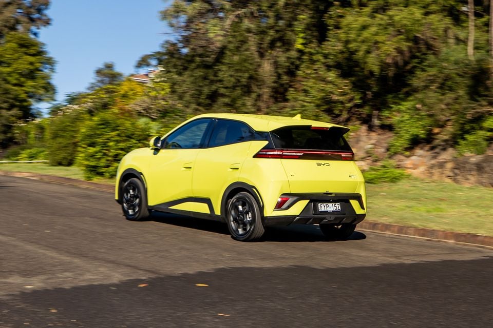 A yellow-green 2026 BYD Atto 1 SUV seen from the rear three-quarter angle while driving on a road surrounded by trees