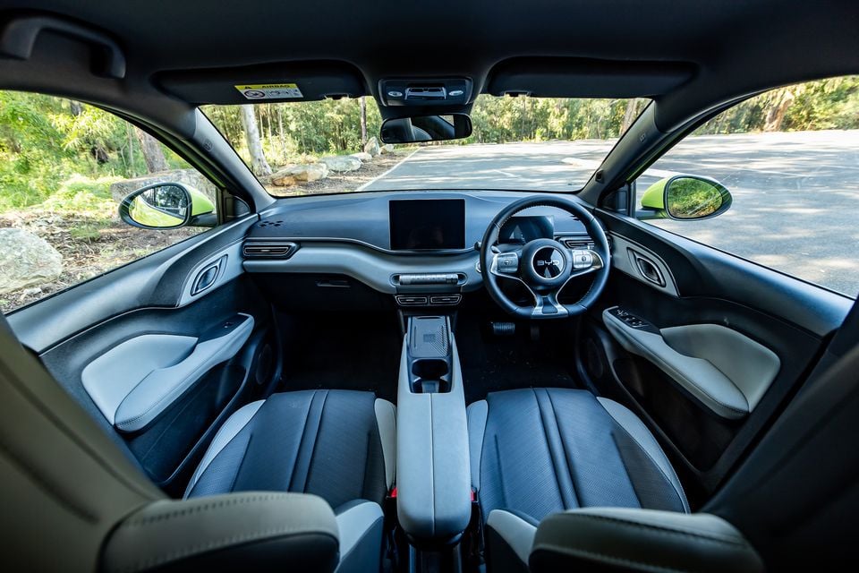 Interior of a BYD Atto 1 showing the dashboard, steering wheel, front seats and centre console, with a touchscreen infotainment display, viewed from the rear cabin