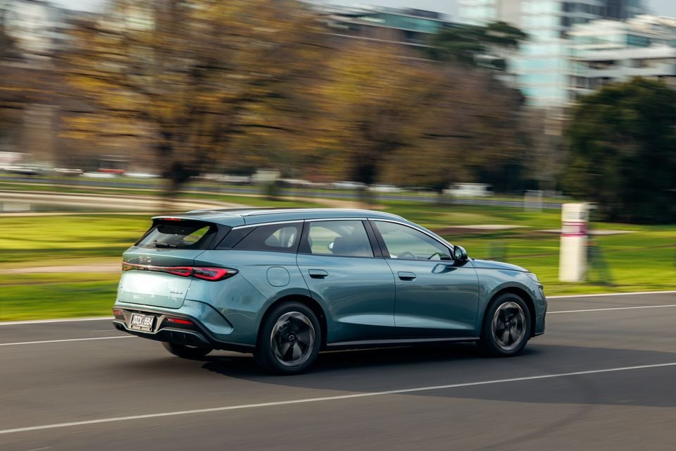 A teal BYD Seal 6 Touring wagon seen from the rear three-quarter angle while driving on a road, with trees and buildings blurred in the background.