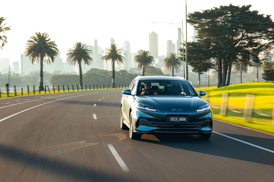 A teal BYD Seal 6 Touring driving along a road with palm trees and a city skyline in the background, seen from the front