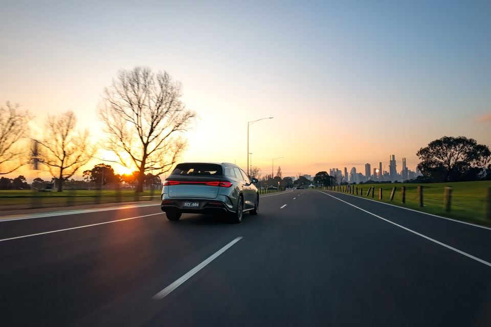 A light blue 2026 BYD Seal 6 Touring SUV seen from the rear while driving on a road at sunset, with a city skyline visible in the distance