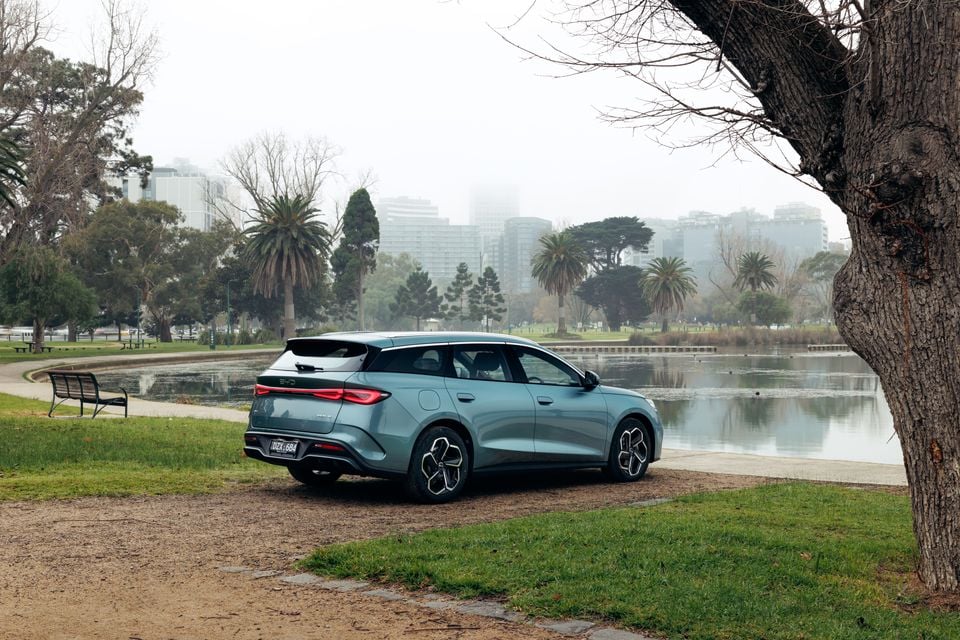 A blue-grey BYD Seal 6 Touring wagon seen from the rear three-quarter angle, parked beside a lake in a park with misty city buildings in the background.