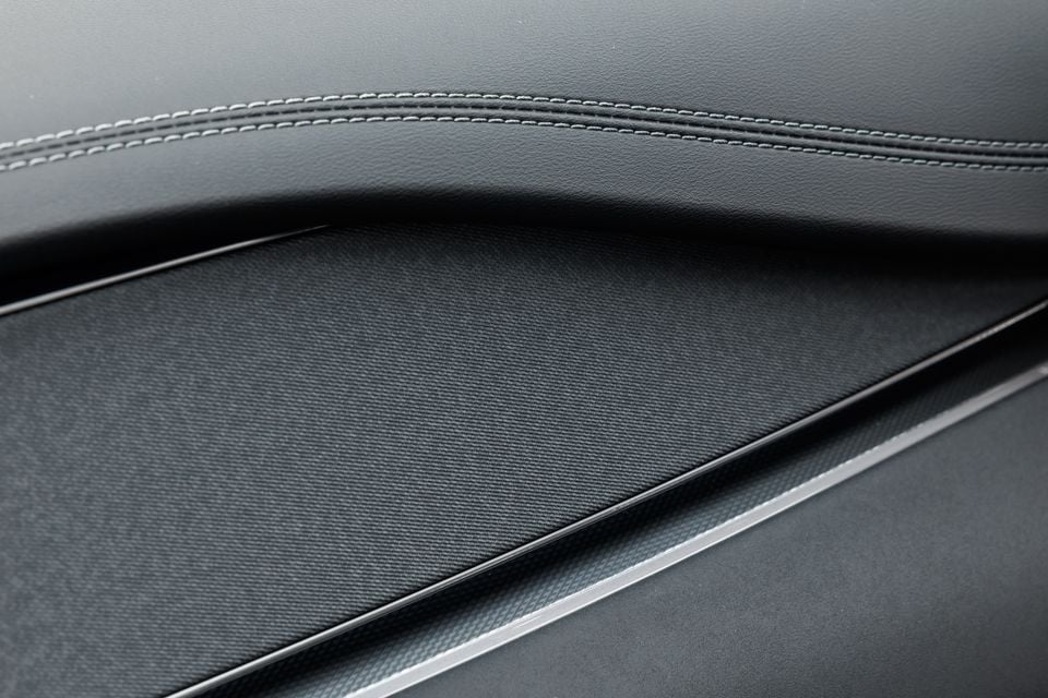 Close-up of a car door panel showing dark leather upholstery with contrast stitching, fabric insert, and a chrome trim strip
