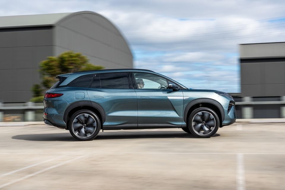 A teal 2026 BYD Sealion 8 SUV seen from the side while driving past industrial buildings