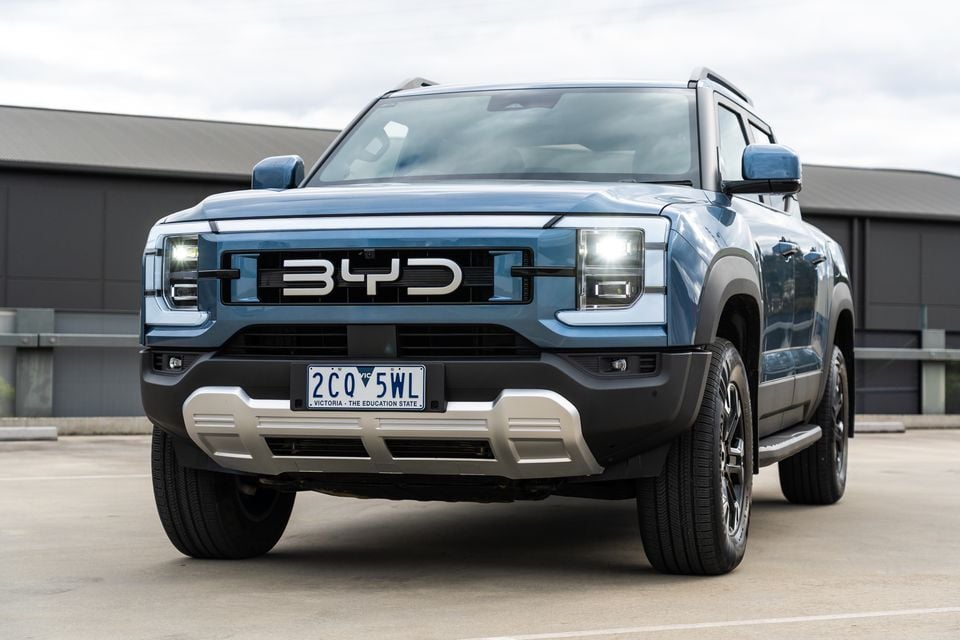 A blue 2026 BYD Shark 6 Premium dual-cab ute seen from the front with its headlights on, parked on a rooftop car park
