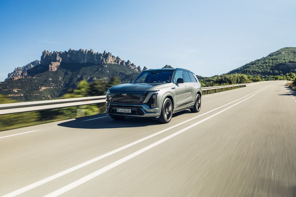 A silver 2026 Cadillac Vistiq SUV driving on a highway with a rocky mountain landscape in the background