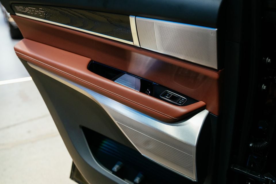 Close-up of the interior door panel of a 2026 Cadillac VISTIQ Platinum, showing tan leather armrest with white stitching, brushed aluminium trim, speaker grilles, and window controls