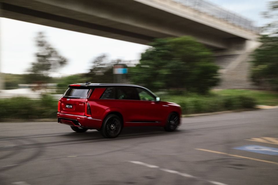 A red 2026 Cadillac Vistiq large SUV captured in motion from the rear three-quarter angle, driving under a freeway overpass