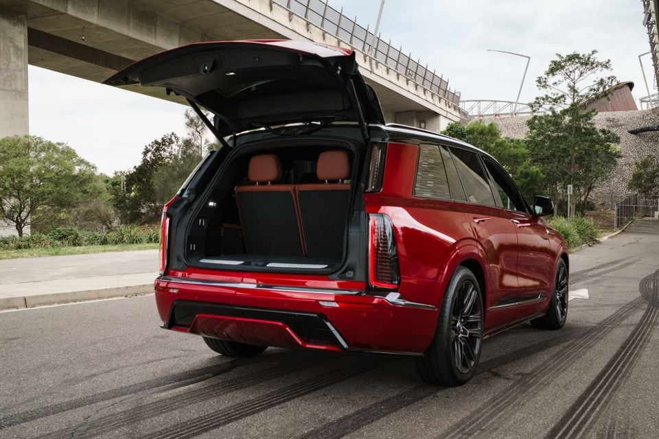 A red 2026 Cadillac XT6 large SUV seen from the rear with the tailgate open, revealing the cargo area and brown leather rear seats, parked on a street under an overpass
