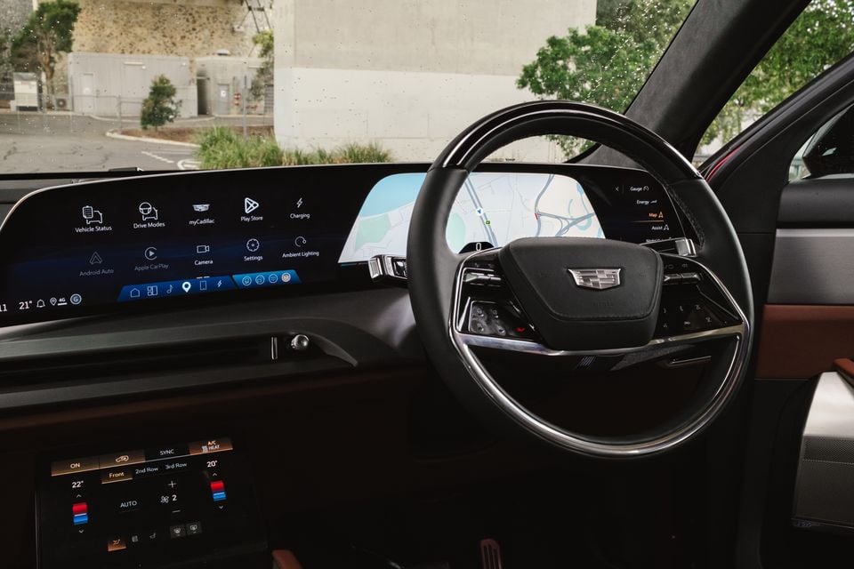 Cadillac VISTIQ interior showing the large curved infotainment screen with Android Auto, Apple CarPlay and charging menus, digital instrument cluster displaying a map, and the Cadillac-badged steering wheel with climate controls below
