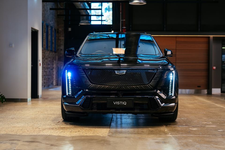 A black Cadillac Vistiq SUV seen from the front, parked inside a modern showroom with LED headlights illuminated