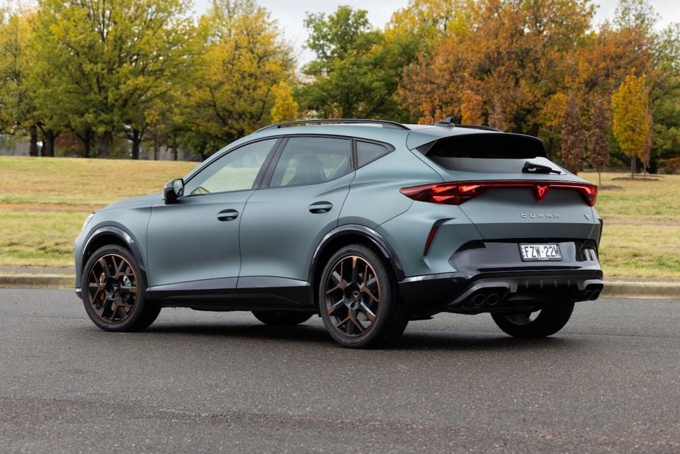 A matte grey-green 2026 Cupra Formentor VZx SUV seen from the rear three-quarter angle, parked on a road with autumn trees in the background