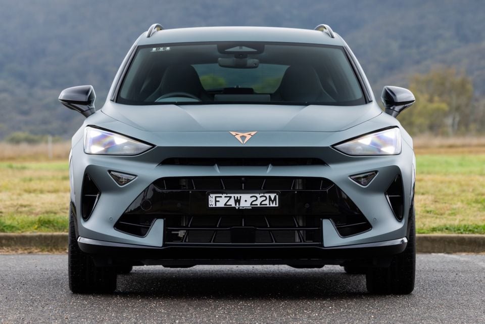 A grey 2026 Cupra Formentor VZX SUV seen from the front, with headlights on, parked on a road with a hilly backdrop