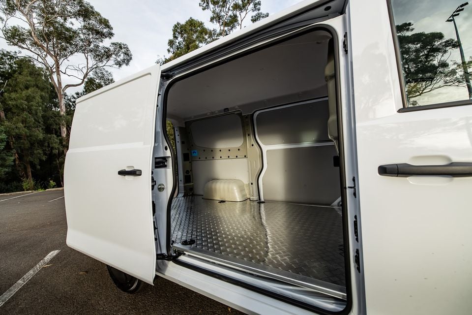 Interior cargo area of a white van with sliding side door open, showing a metal chequer-plate floor and bare panel-lined walls, parked outdoors