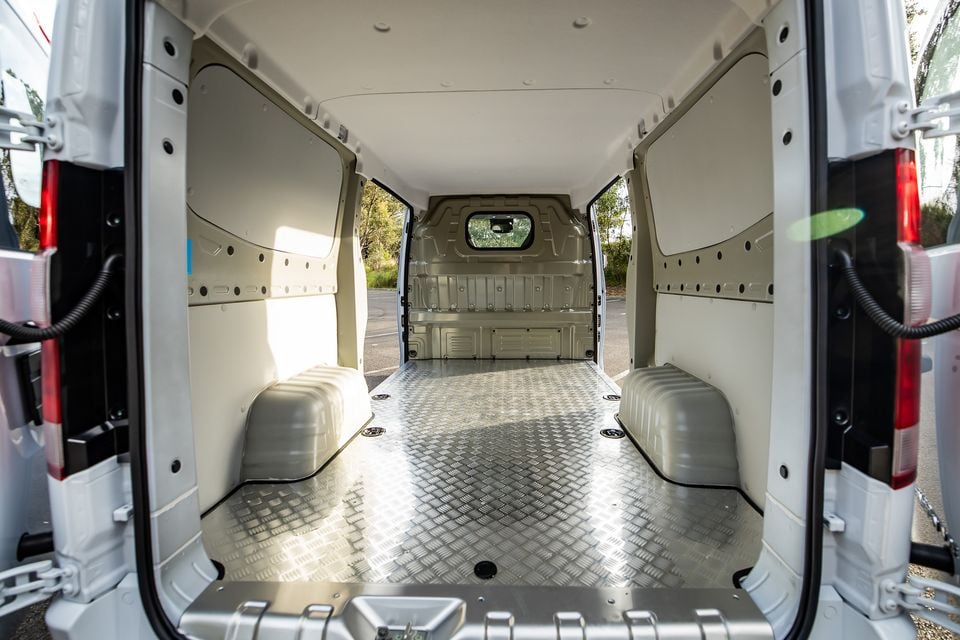 The empty cargo area of a van with a chequer-plate aluminium floor and white lined walls, viewed from the open rear doors