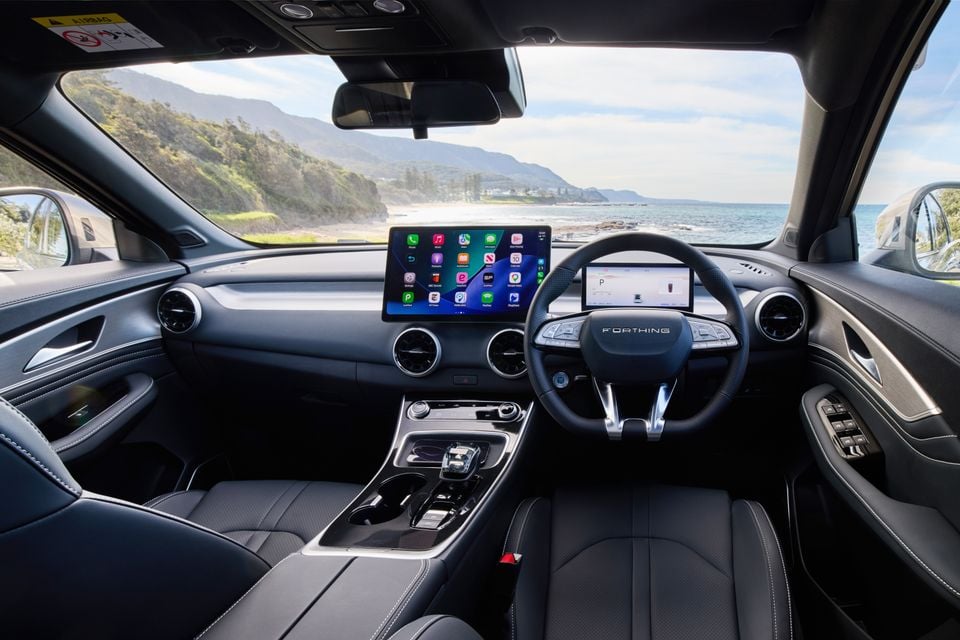 Interior of a Forthing Taikon 5 showing the dashboard, large central touchscreen displaying Apple CarPlay, digital instrument cluster, and steering wheel with coastal scenery visible through the windscreen