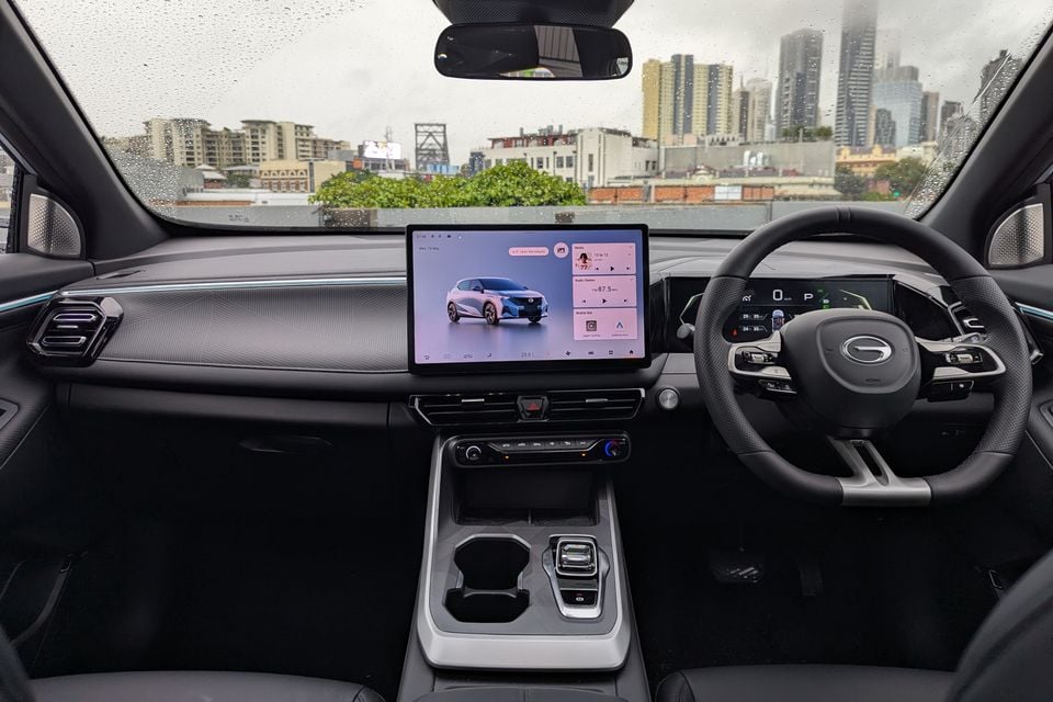 Interior view of a GAC Emzoom showing the dashboard, large central touchscreen infotainment display, steering wheel, digital instrument cluster, and centre console, with a city skyline visible through the windscreen