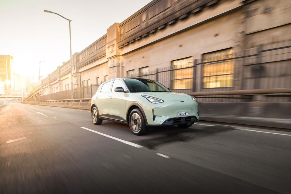 A light green 2026 Geely EX2 Inspire small SUV driving on a city road at golden hour, seen from the front side