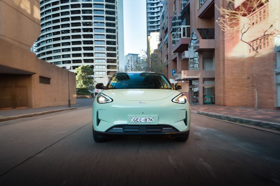 A light green 2026 Geely EX2 Inspire SUV driving through a city street, seen head-on with headlights on and urban buildings in the background